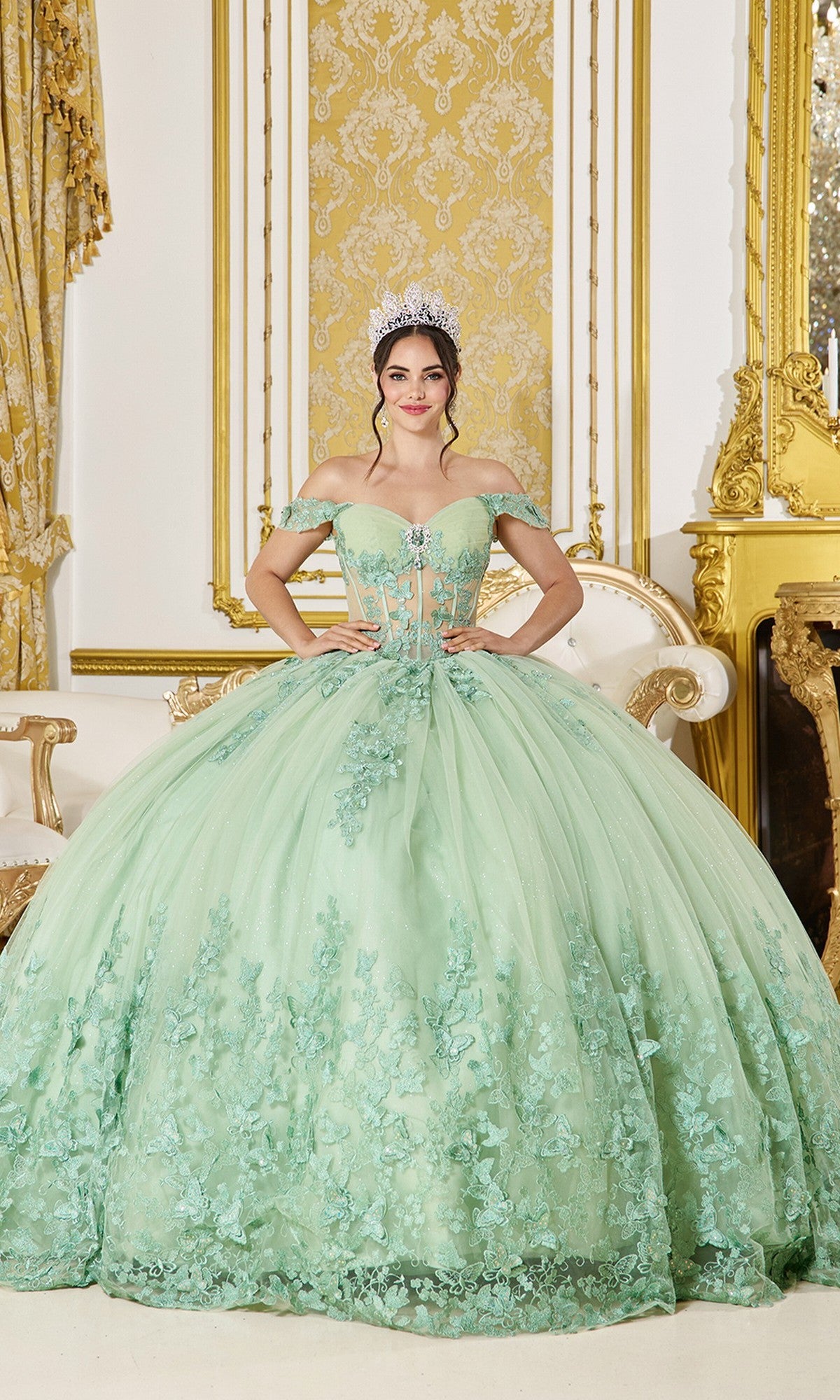 A young woman stands in an ornate room, wearing the Sage Green Quinceanera Dress 5065 by Dancing Queen with floral embroidery and 3D butterfly appliqué, complemented by a sparkling tiara.